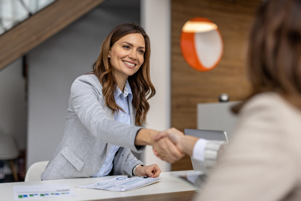 Two businesswomen shaking hands in a modern office, expressing joy and satisfaction after sealing a successful deal. Smiling faces reflect the strong partnership and collaboration achieved